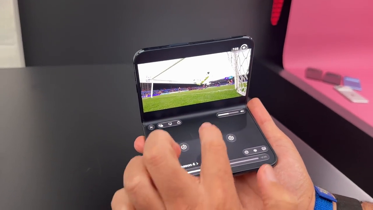 iPhone Duo folded halfway on a table showing a video playing on the top screen and media controls on the bottom screen